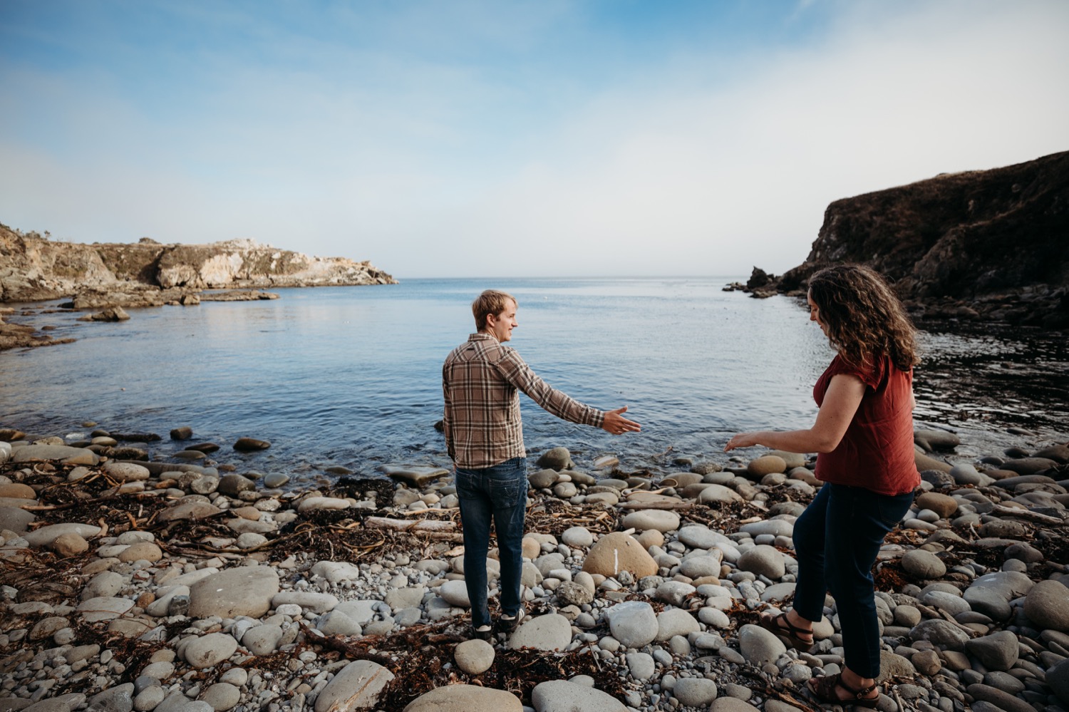 Man reaches his hand out to help his fiance across a rocky beach in Salt Point Park during their engagement photoshoot.