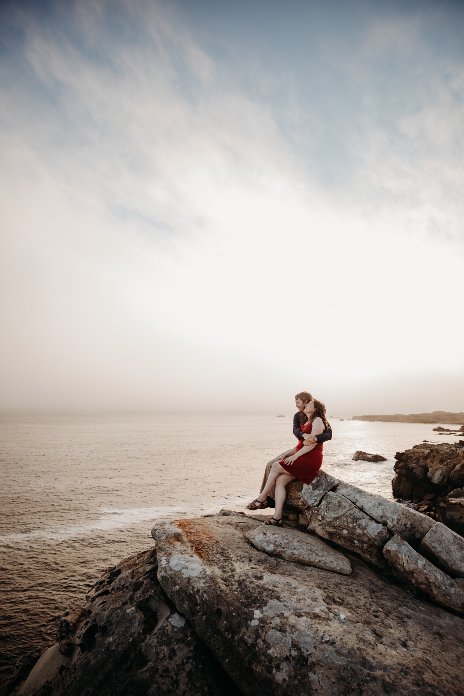 Man holds his fiance while they sit on a cliff at sunset in Salt Point State Park during their engagement photoshoot.