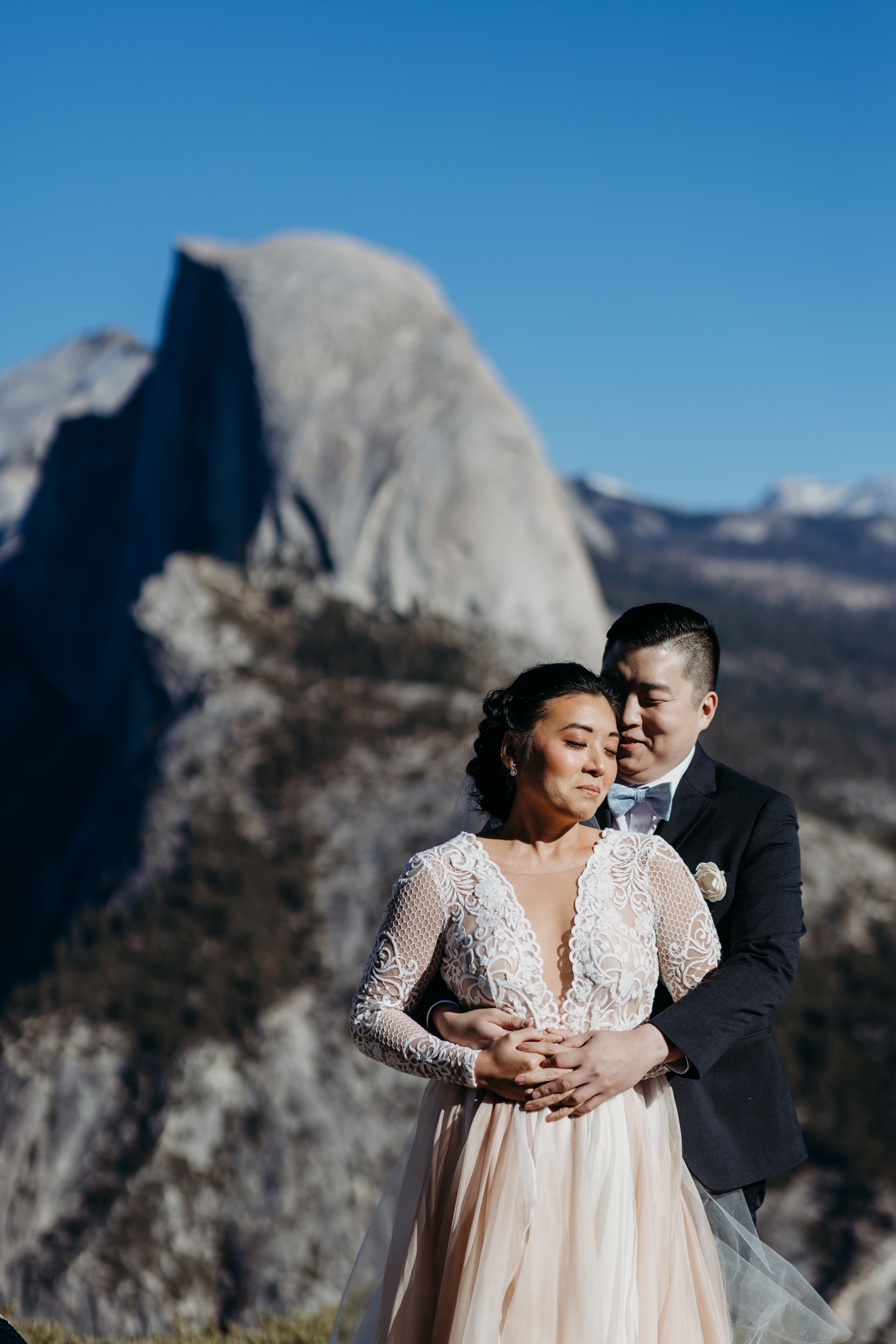 Groom embraces his bride from behind with Yosemite's Half Dome in the background.