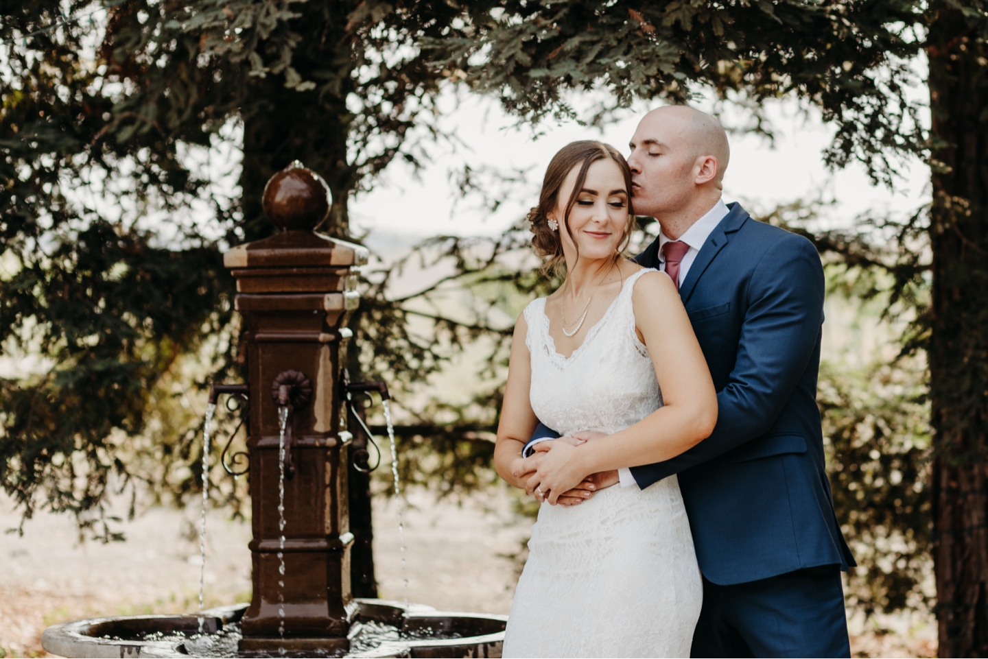 Groom hugs bride from behind and kisses her forehead as she leans into him and smiles. Wedding photography in Sacramento by Liz Koston.