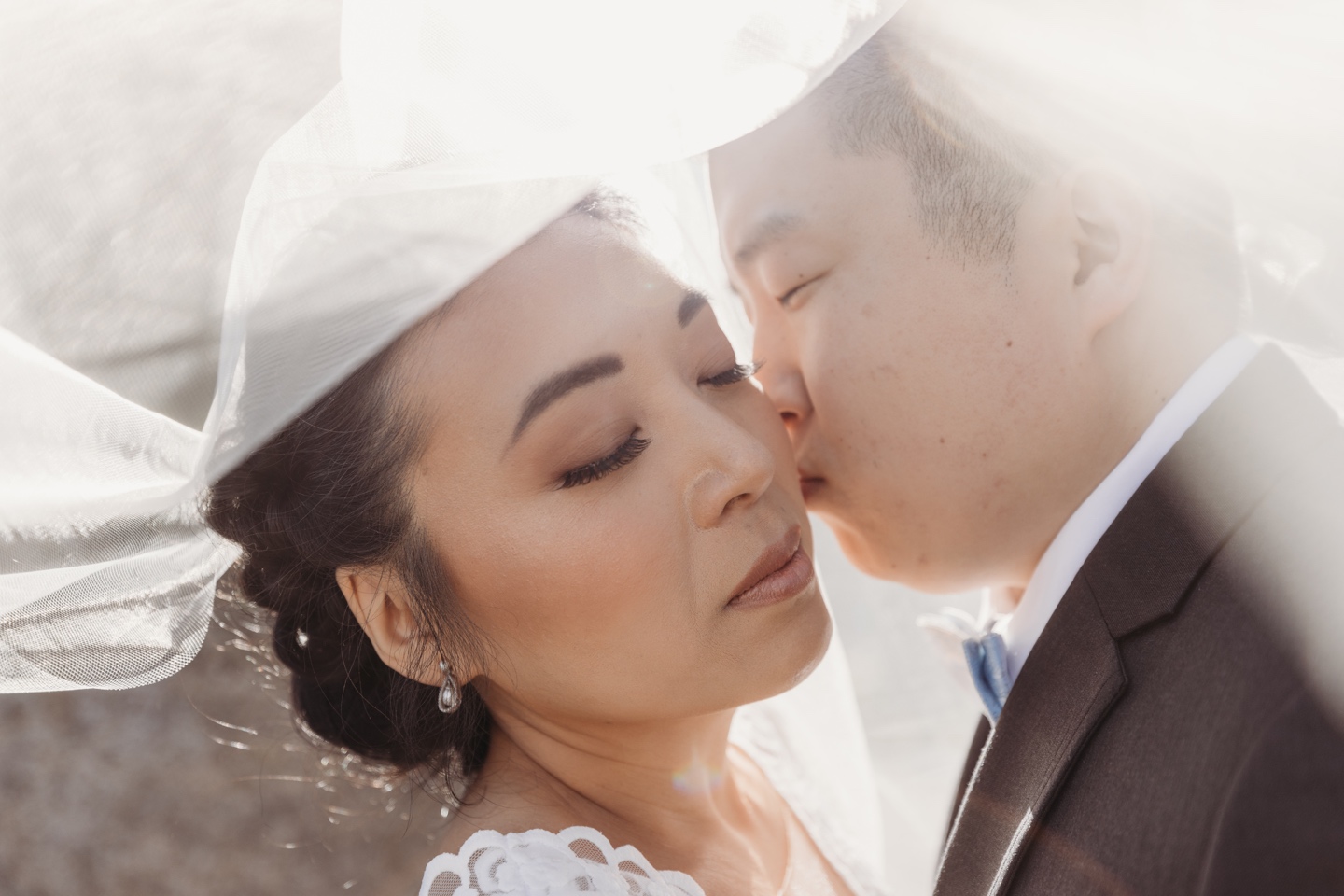 Groom kisses bride on the cheek as they're surrounded her veil and an ethereal glow.
