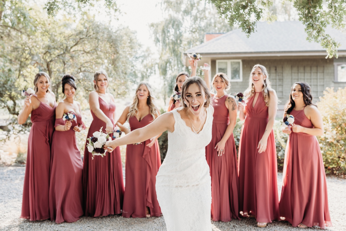 Bride stands and smiles big in front of her bridesmaids who all smile at her in their dark pink bridesmaids dresses. Wedding photography in Sacramento by Liz Koston.