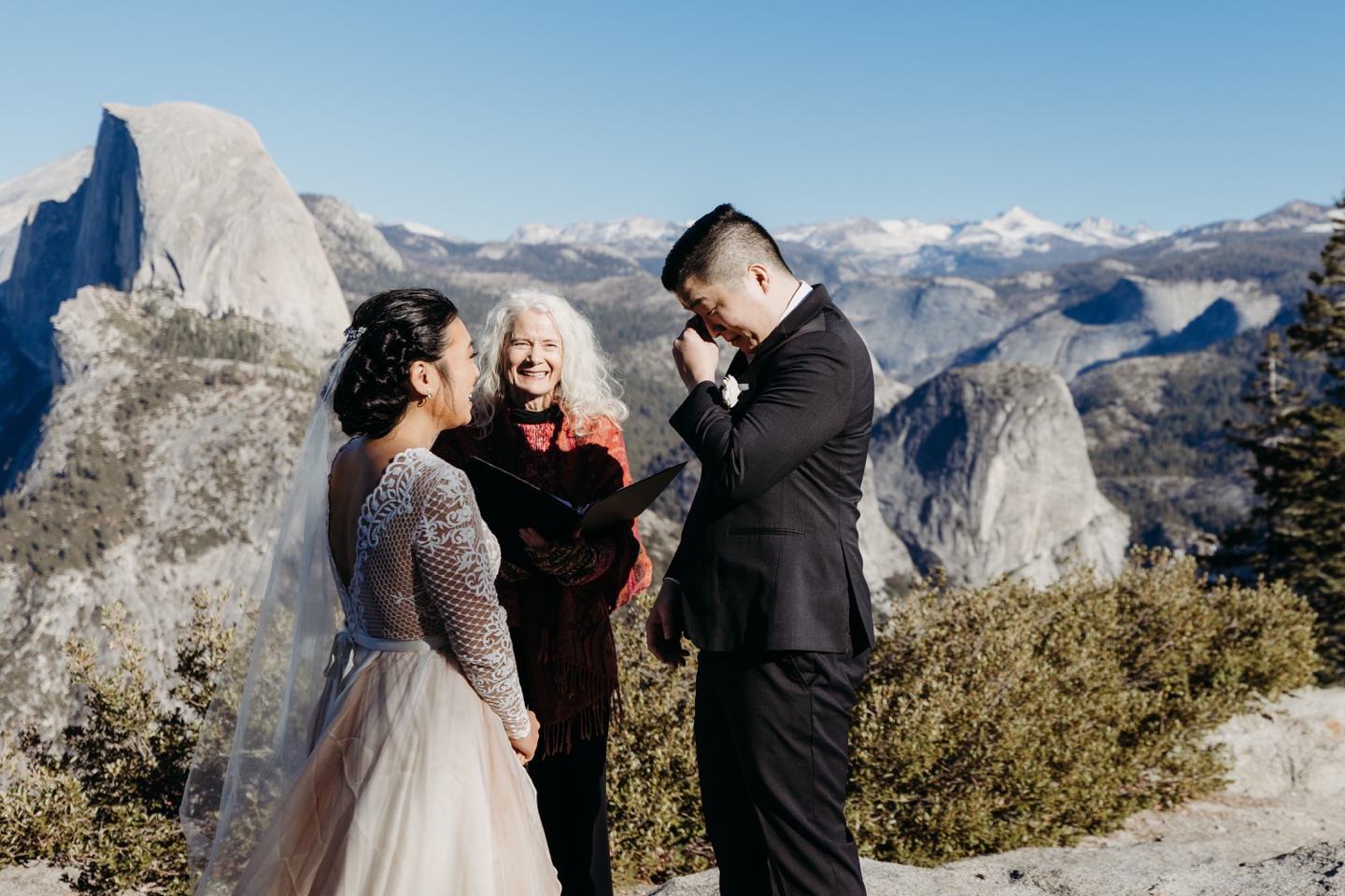 Groom dries his eyes as he says his vows and the bride and officiant smile at him. There are views of Yosemite in the distance. Photos taken by Yosemite elopement photographer, Liz Koston.