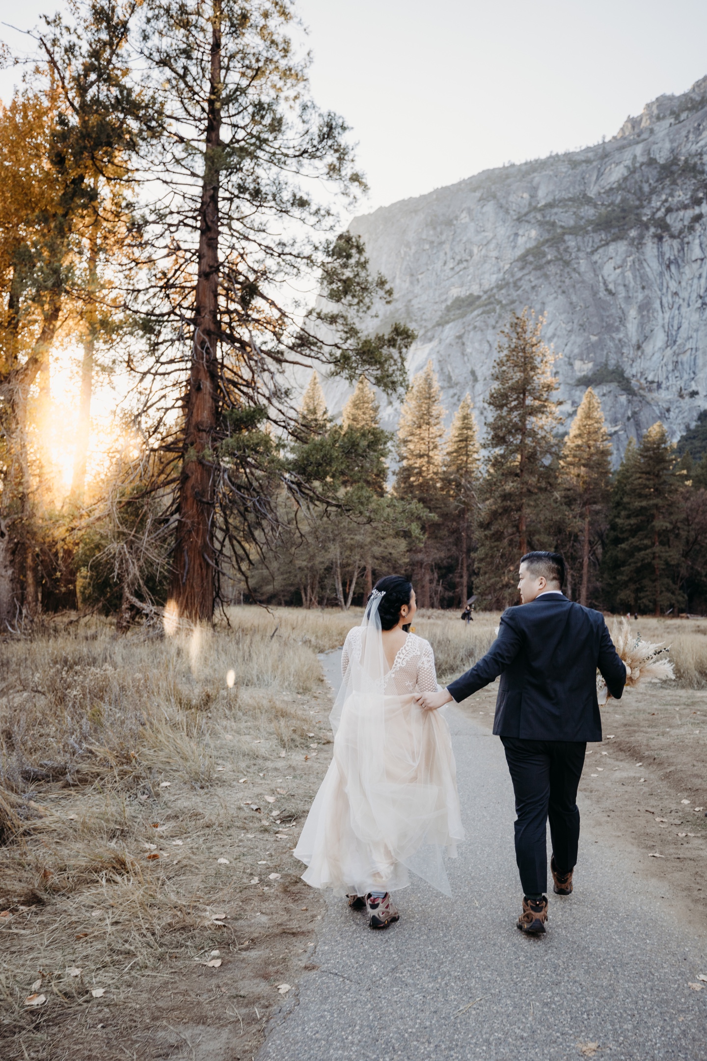 Groom helps his bride hold her wedding dress as they walk down a hiking trail in Yosemite as the sun sets after their elopement.