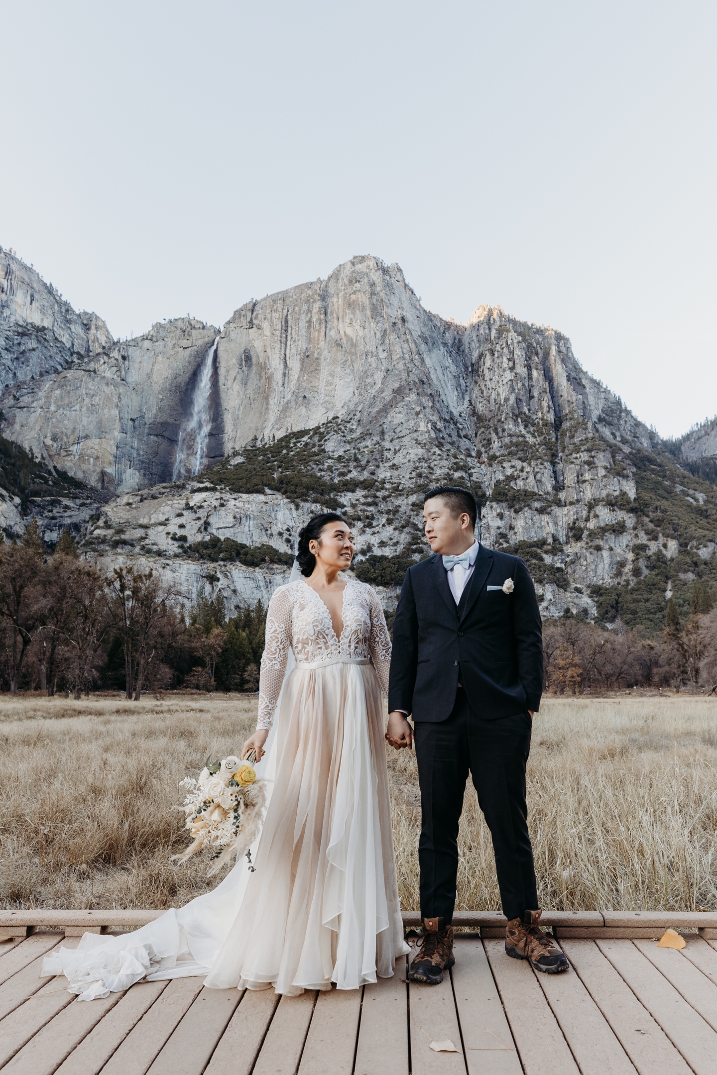 Bride and groom hold hands on a wood walking path in front of Yosemite Falls after their Yosemite wedding.