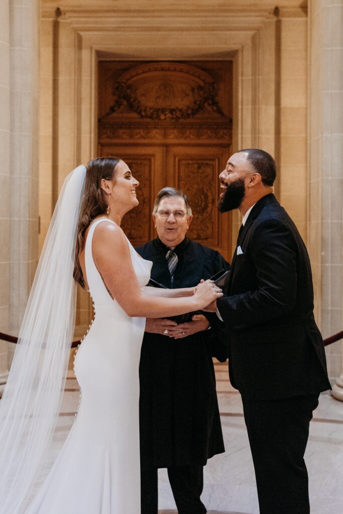 Bride and groom hold hands and smile wide at each other as the officiant performs their civil ceremony during their San Francisco City Hall wedding. Photo by Liz Koston Photography.