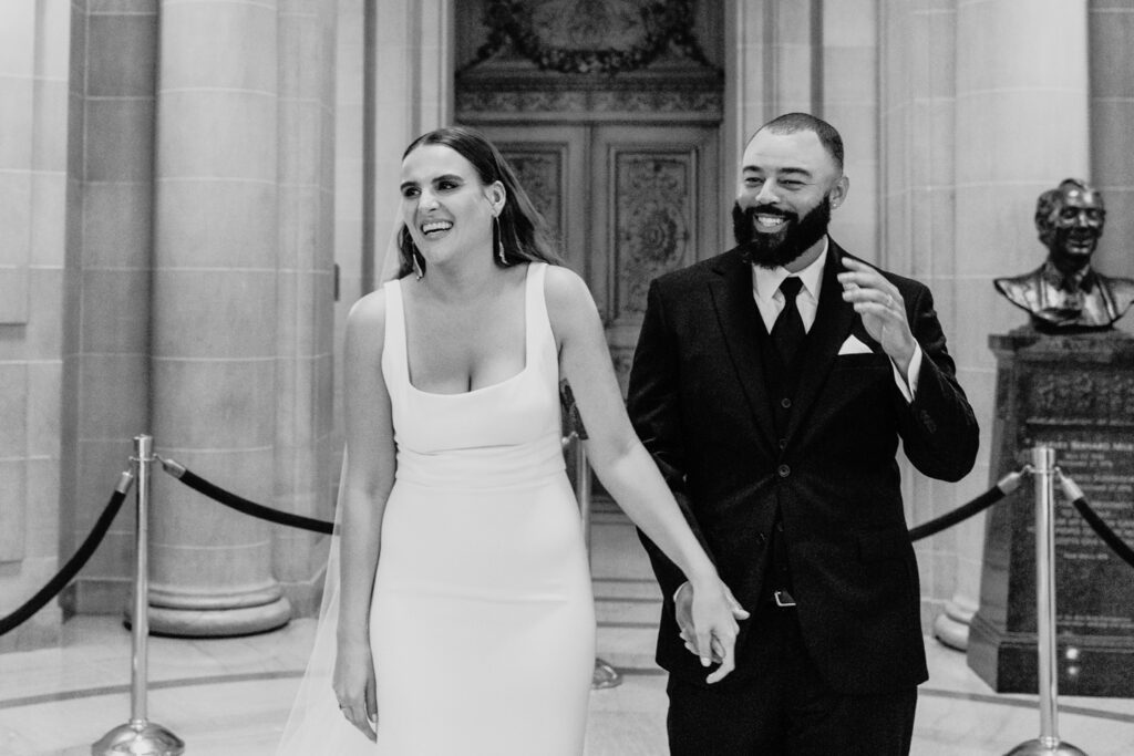 Bride and groom smile widely while holding hands as they walk to elope in San Francisco City Hall. Photo by Liz Koston Photography.