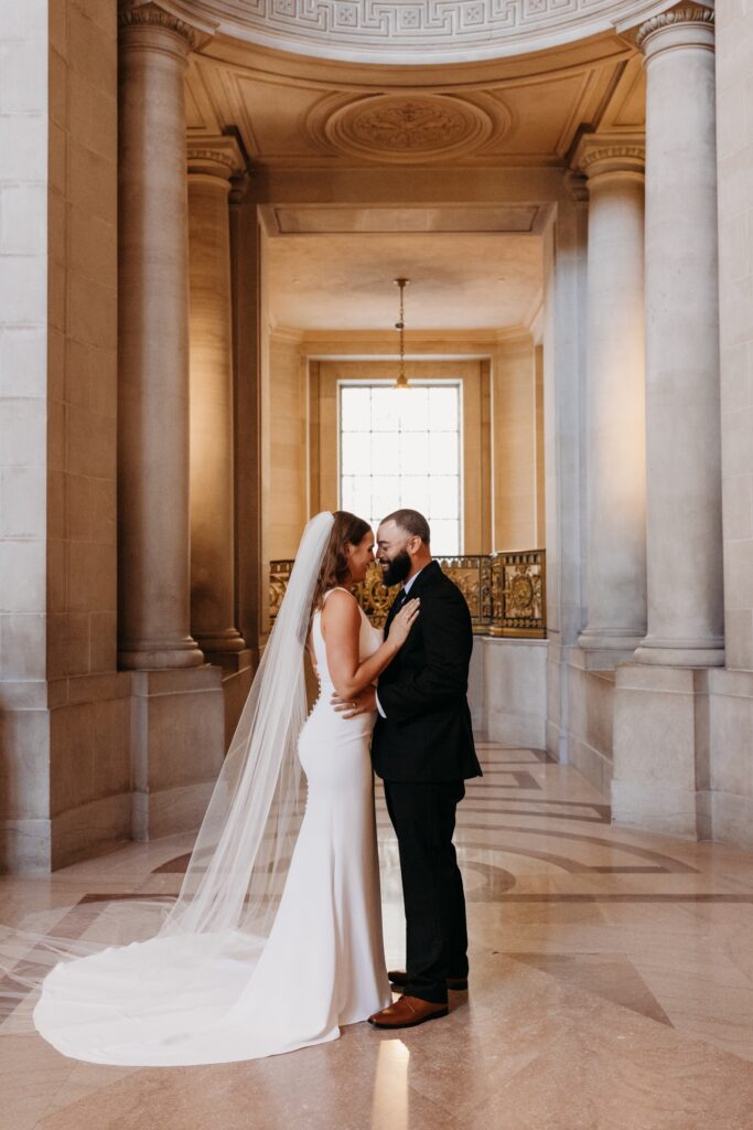 Bride and groom stand embracing each other, touching noses and smiling in San Francisco City Hall. Photo by Liz Koston Photography.