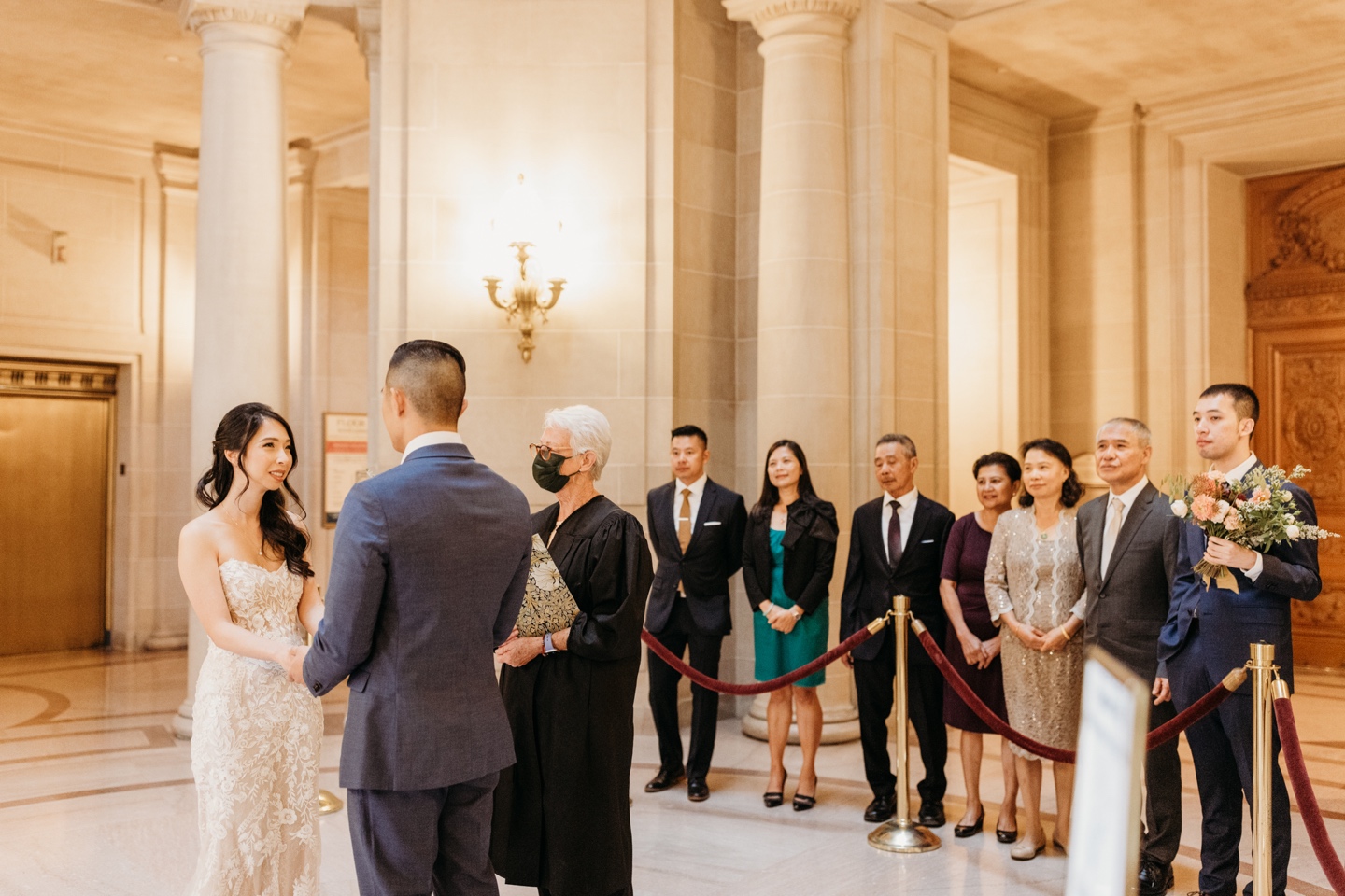 Bride and groom hold hands while exchanging vows and their family and friends watch during their San Francisco City Hall elopement ceremony.