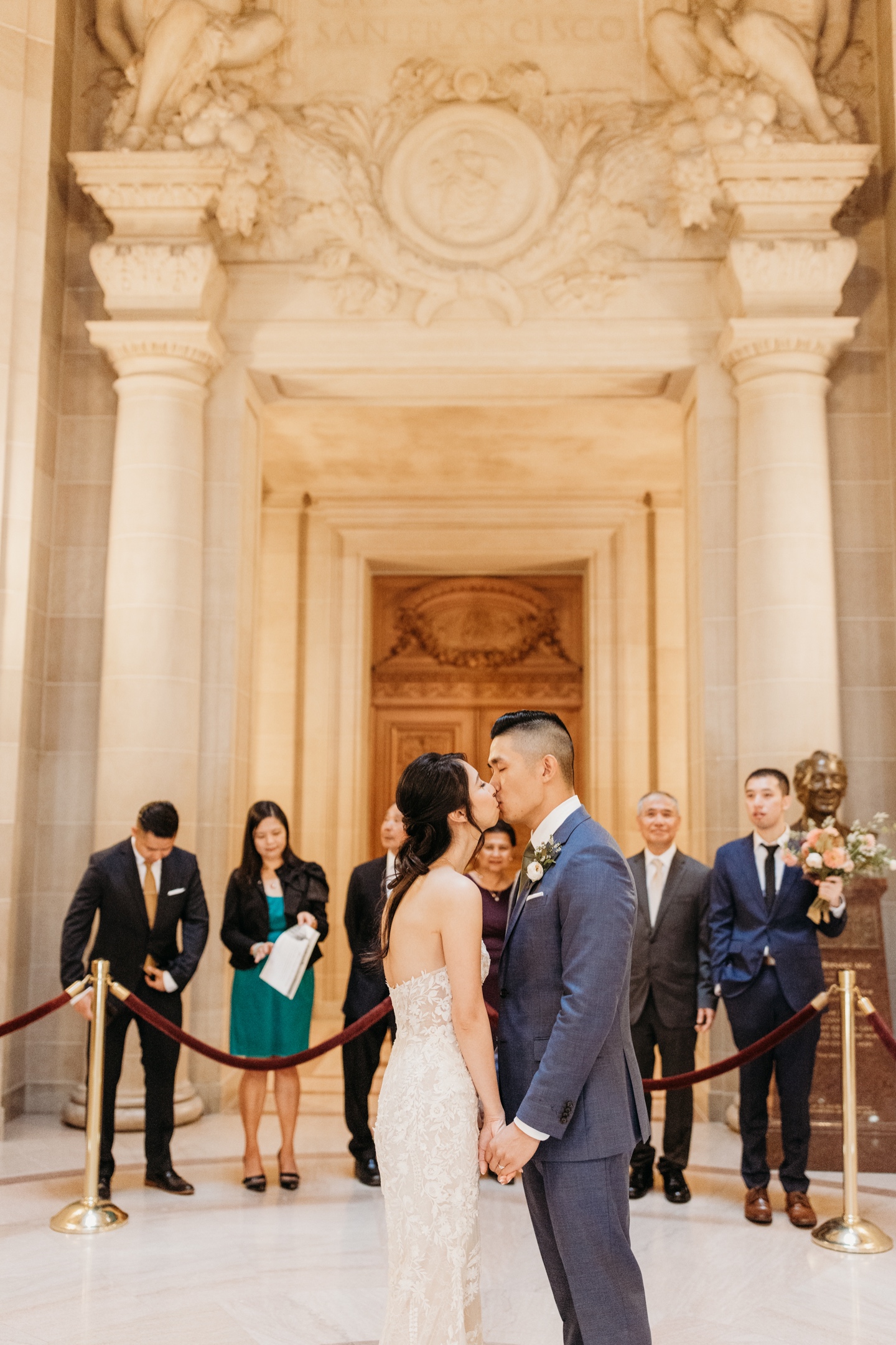 Bride and groom kiss after exchanging vows during their San Francisco City Hall elopement ceremony while their family watches.
