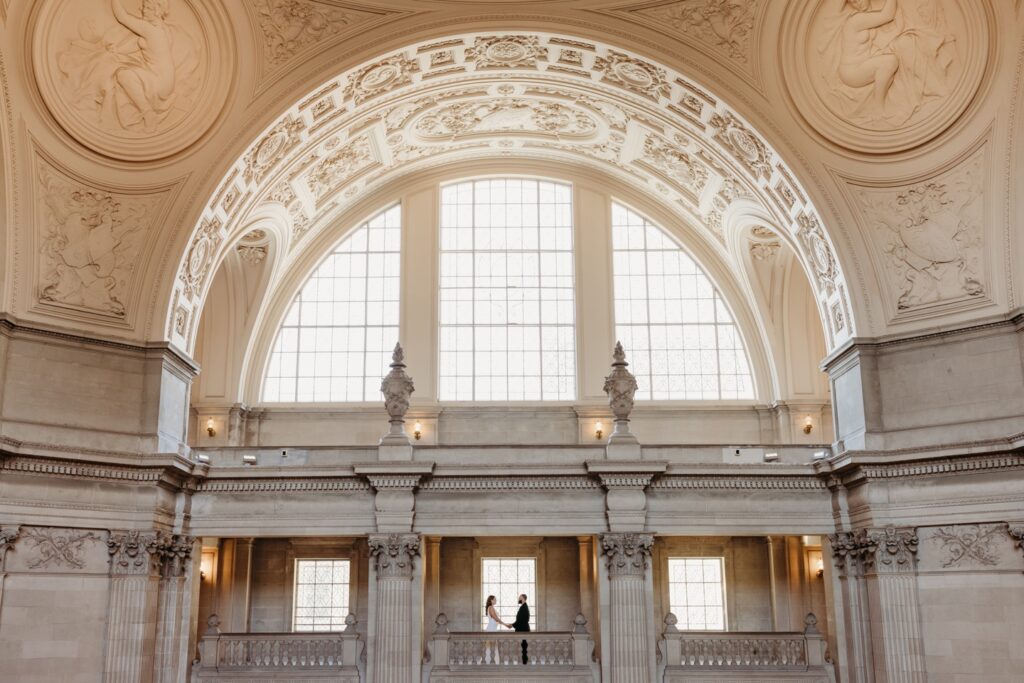 Bride and groom stand holding hands underneath the beautiful San Francisco City Hall ceiling. Photo by Liz Koston Photography.