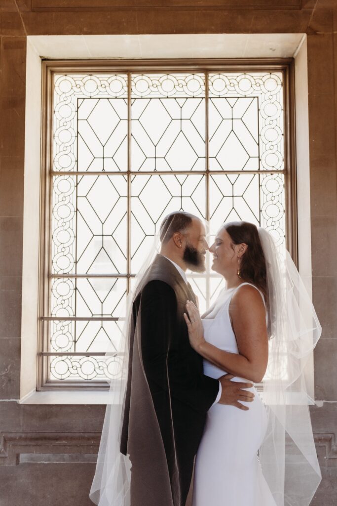 Bride and groom touch noses with brides veil covering them both in front of a window during their San Francisco City Hall wedding. Photo by Liz Koston Photography.