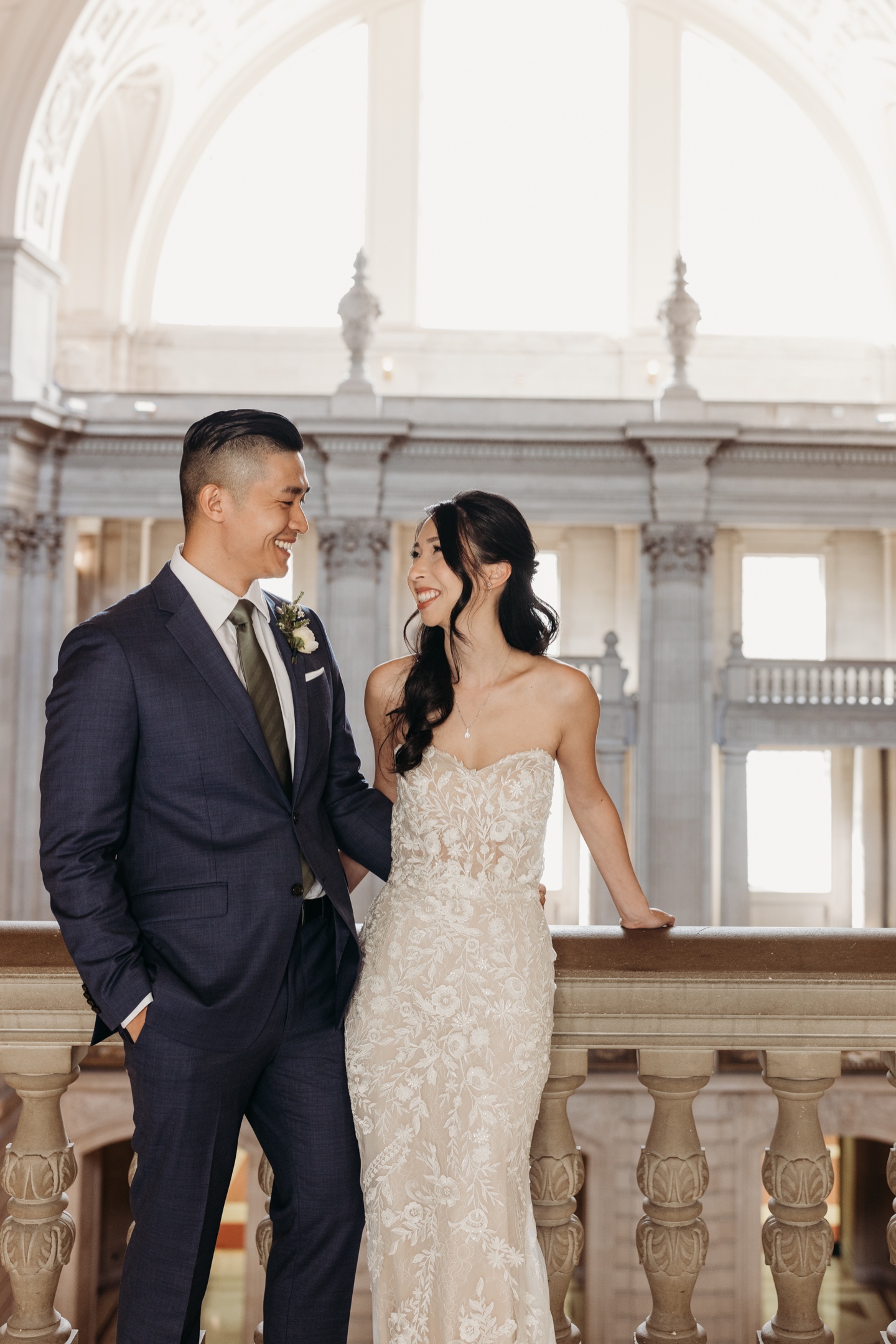 Bride and groom sand side by side smiling at each other in a San Francisco City Hall balcony.
