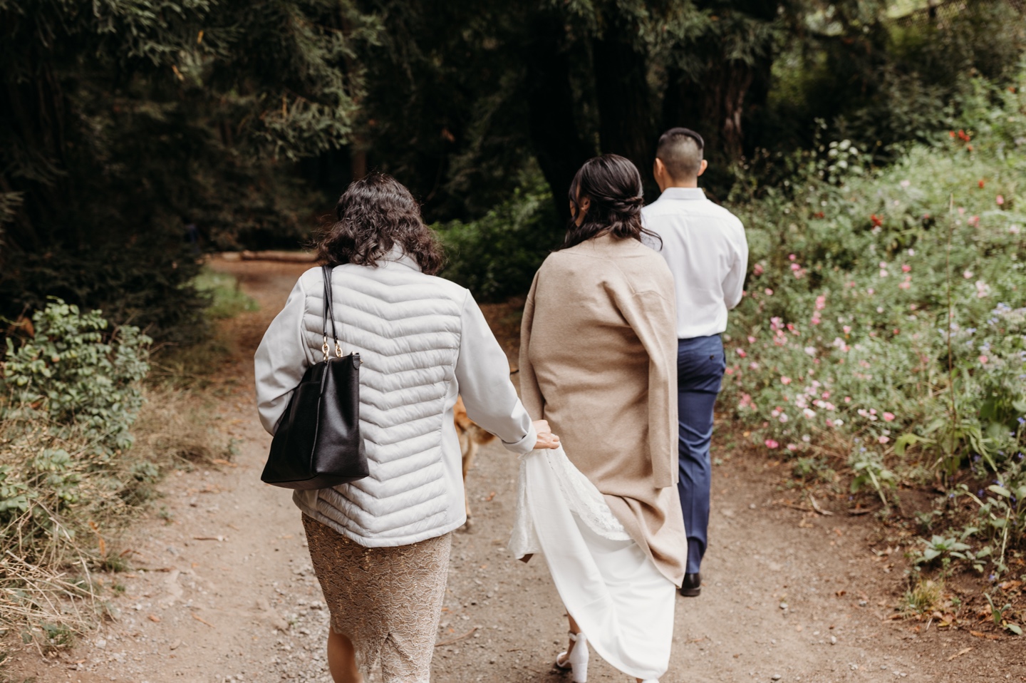 Bride and groom walk through forest path while Mother of the Bride holds the bride's dress train.