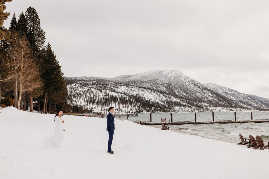 Bride walks up behind her groom for a private first look on the shores of Lake Tahoe with the snowy mountains in the distance. Liz Koston Photography.