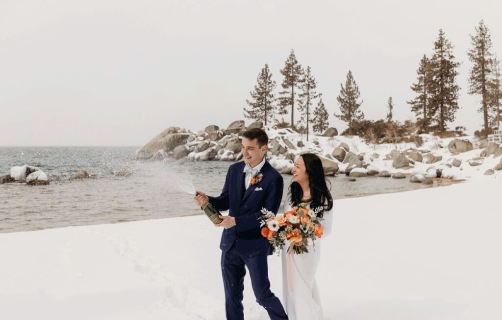 Groom pops a champagne bottle as he and his bride holding an orange bouquet of flowers walk along the shores of Lake Tahoe in the snow. Liz Koston Photography.