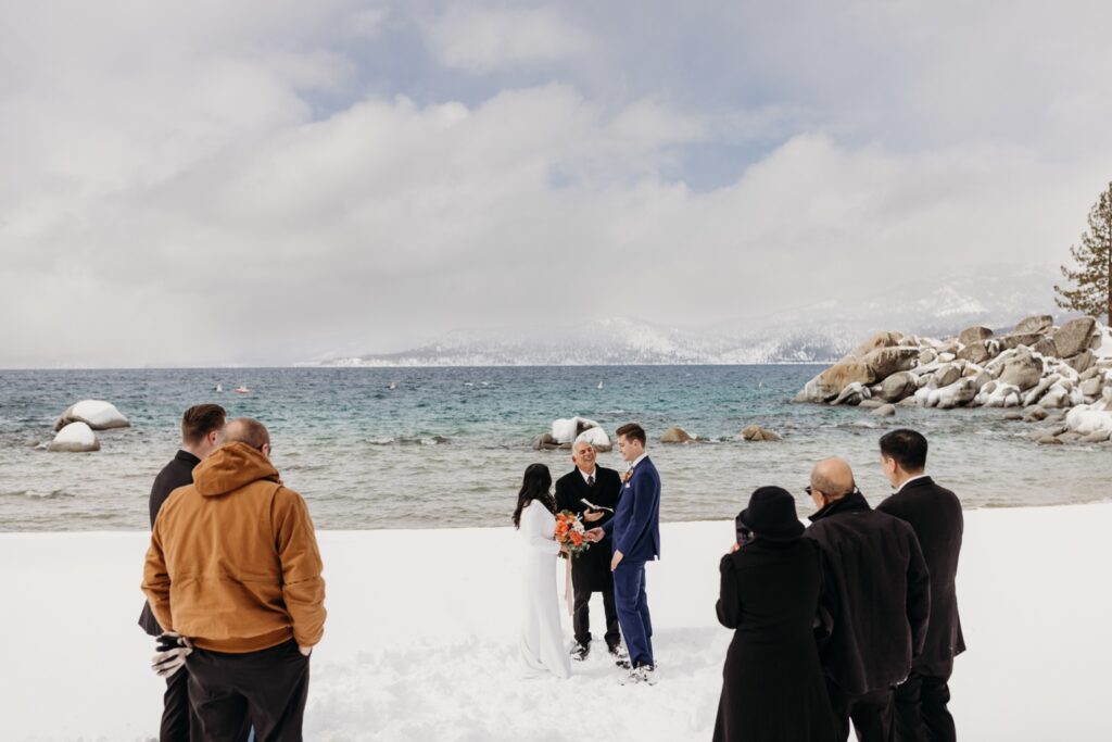 Guests watch the bride and groom exchange vows on the shores of Lake Tahoe as the sun breaks through the clouds. Liz Koston Photography.