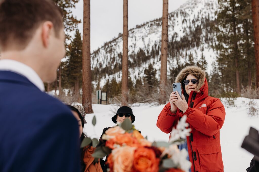 Guest wearing a puffy red winter coat takes a photo of the couple with her phone. Liz Koston Photography.