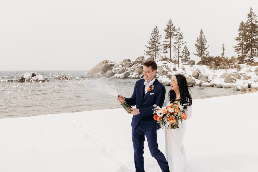 Groom pops champagne as his bride walks beside him holding her bouquet of flowers on the snowy banks of Lake Tahoe. Liz Koston Photography.